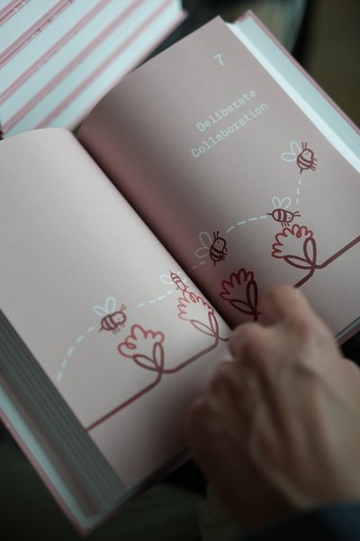 A hand holding open the Deliberate Collaboration chapter, showing bees and flowers drawn in one continuous red line