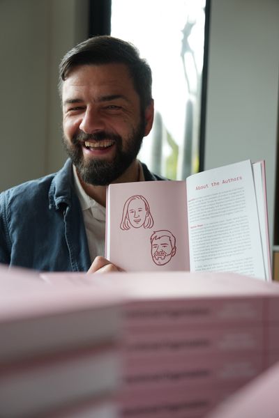 Mathias smiling and holding the book open to the About the Authors page, which shows small illustrated portraits of Sara and Mathias